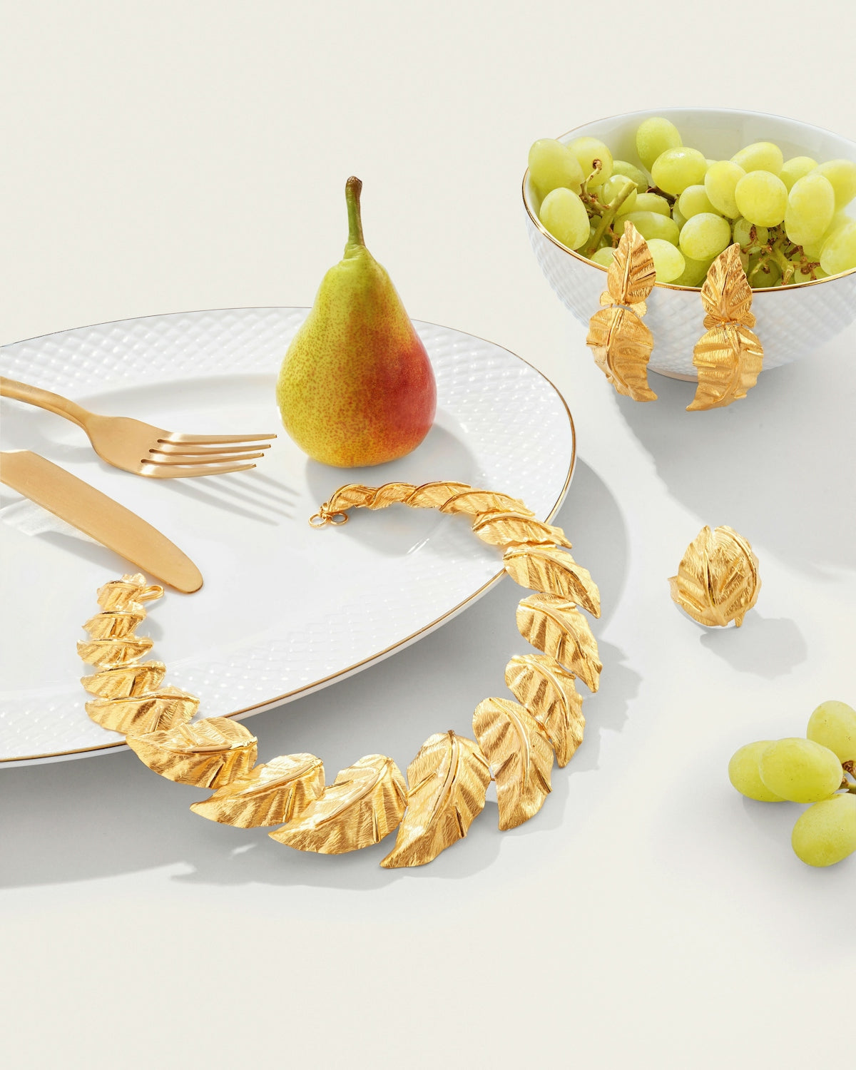 Gold leaf jewelry, pear, grapes, and cutlery on white.